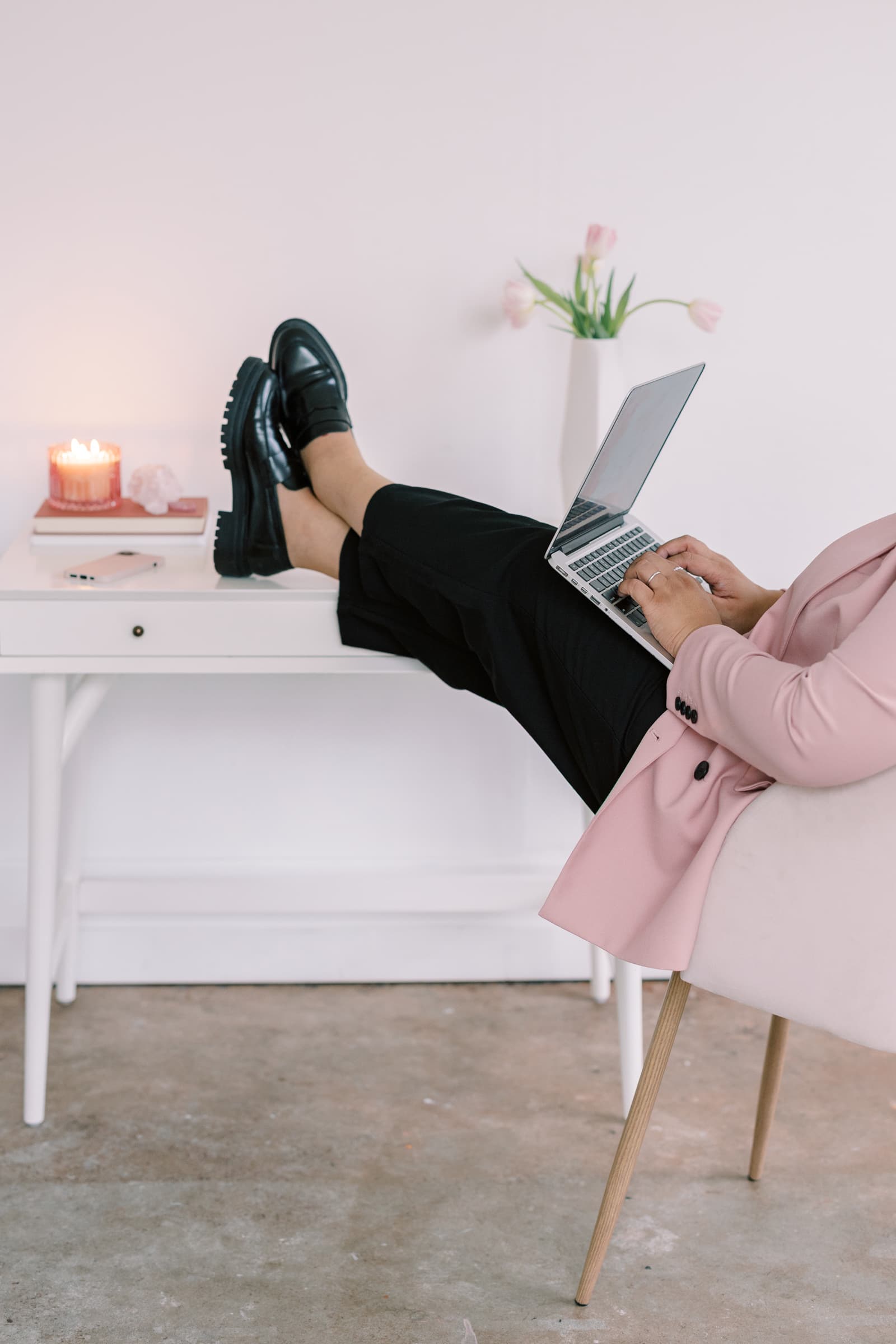 A woman in a pink blazer with black pants and shoes kicks her feet up while working on their laptop.